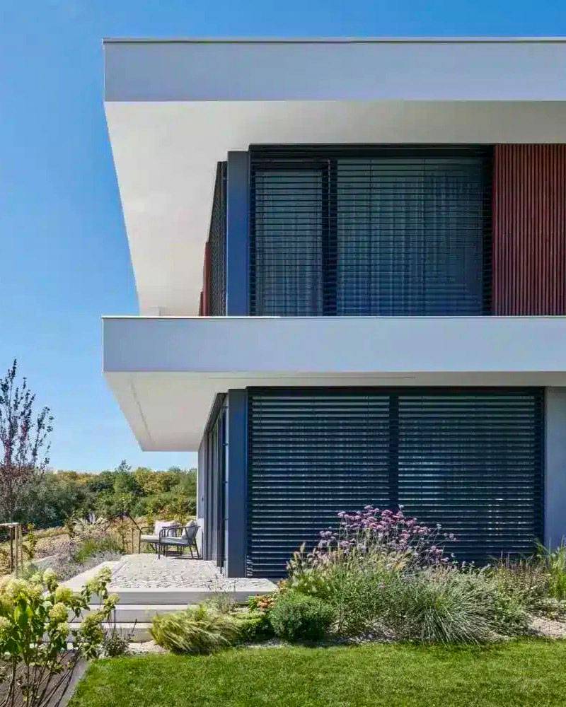 Moderná dvojpodlažná rodinná vila s vonkajšími čiernymi žalúziami na veľkých presklených oknách, minimalistickou architektúrou, bielou fasádou a upravenou záhradou v slnečný deň.